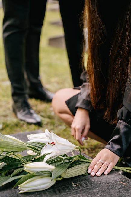 A woman dressed in black kneels at a grave, arranging a floral tribute of white lilies and greenery on a dark stone surface. The lilies are fresh, with open petals and long green stems, complemented b
