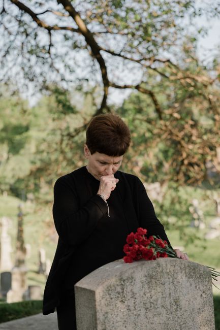 A woman with short dark hair, dressed in black, stands in a cemetery under a tree with green and orange leaves. She holds a bouquet of vibrant red carnations and appears to be in a moment of reflectio