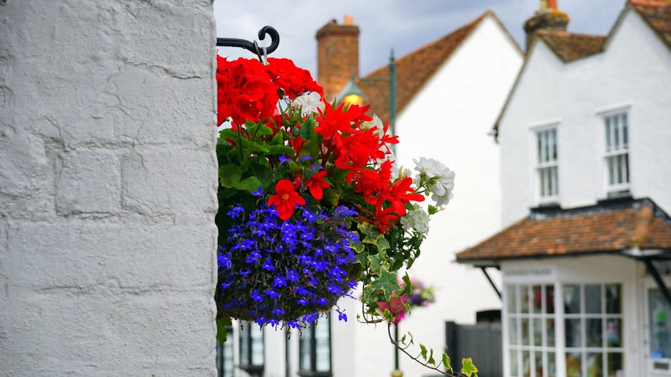 A vibrant hanging flower basket affixed to a white brick wall, featuring a mix of red geraniums, white petunias, and purple lobelias. The flowers are lush and fresh, with full petals displaying bright