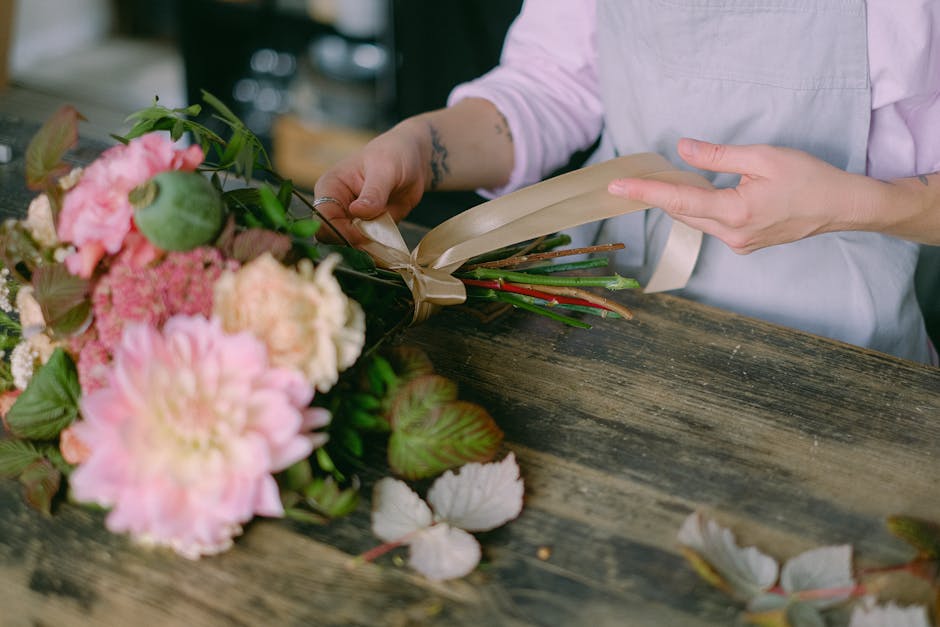 A person in a light pink shirt and white apron is wrapping a bouquet of fresh flowers on a rustic wooden table in a floral workshop. The bouquet features pink peonies, green leaves, and other pink and