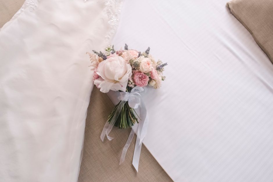 A delicate bouquet of fresh flowers lies on a white bedspread, arranged with soft pink roses, white peonies, and lavender sprigs. The bouquet is wrapped in semi-transparent white tissue paper and tied