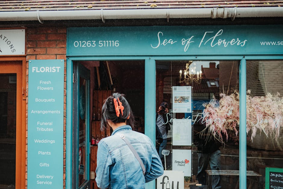 Front view of the florist shop 'Sea of Flowers' on West Wickham High Street, with a woman wearing a denim jacket and a large orange hair clip looking into the shop window. Through the glass, the inter